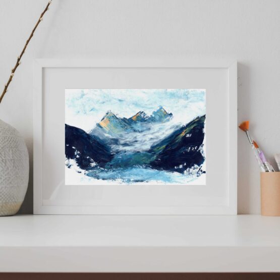 A framed Landscape painting witrh blue mountains capped with orange snowy peaks rests on a grey shelf against a grey wall next to a vase with sticks in it and paint brushes
