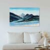 Abstract impressionist landscape painting with blue mountain ranges made of marbled paint with light blue skies and a river below hung on a white wall above a white cushioned couch with light wood accents
