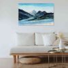 Abstract impressionist landscape painting with blue mountain ranges made of marbled paint with light blue skies and a river below hung on a white wall above a white cushioned couch with light wood accents