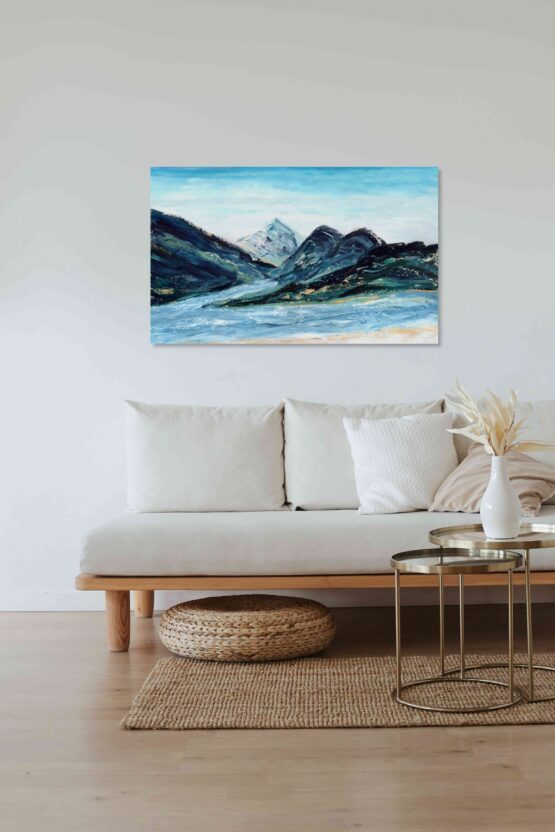 Abstract impressionist landscape painting with blue mountain ranges made of marbled paint with light blue skies and a river below hung on a white wall above a white cushioned couch with light wood accents