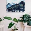 Blue, green and hints of orange and yellow marbled abstract mountain against a wispy white cloudy back ground with bits of blue sky peeking through original painting frameless, hung on a white wall above a white table with a green round leafed plant in a small white plant pot