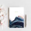 A small journal rests upon a grey counter next to pink and white magnolias and a silver pen. The artwork on the journal is a blue abstract landscape with yellow and orange peeking through. Art is by Melissa Critchlow Fine Artist