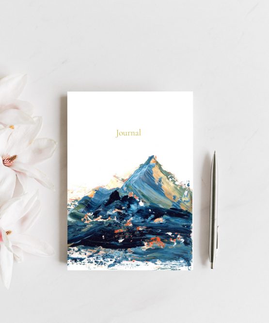 A small journal rests upon a grey counter next to pink and white magnolias and a silver pen. The artwork on the journal is a blue abstract landscape with yellow and orange peeking through. Art is by Melissa Critchlow Fine Artist