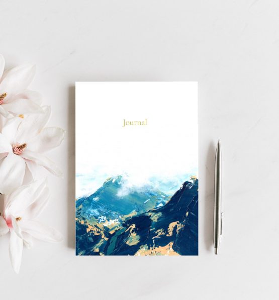 A small journal rests upon a grey counter next to pink and white magnolias and a silver pen. The artwork on the journal is a blue abstract landscape with yellow peeking through. Art is by Melissa Critchlow Fine Artist