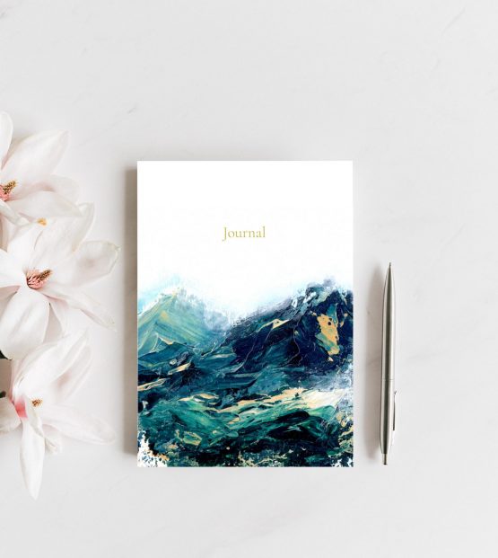 A small journal rests upon a grey counter next to pink and white magnolias and a silver pen. The artwork on the journal is a green and blue abstract landscape with yellow peeking through. Art is by Melissa Critchlow Fine Artist