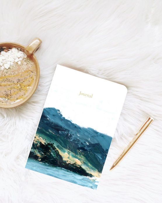 Small journal rests on white fur blanket next to gold pen and a hot drip with white chocolate and chia seeds . The artwork is of a forested Mountain next to a lake by Melissa Critchlow Fine Artist