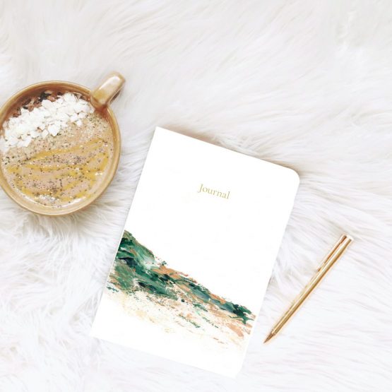 Small journal rests on white fur blanket next to gold pen and a hot drip with white chocolate and chia seeds . The artwork is of an abstract landscape using light brown and green colours by Melissa Critchlow Fine Artist