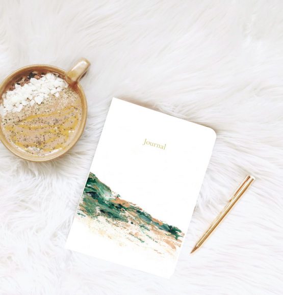 Small journal rests on white fur blanket next to gold pen and a hot drip with white chocolate and chia seeds . The artwork is of an abstract landscape using light brown and green colours by Melissa Critchlow Fine Artist