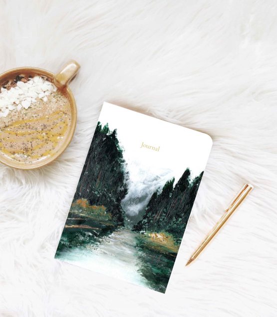 Small journal rests on white fur blanket next to gold pen and a hot drip with white chocolate and chia seeds . The artwork is of a forest by Melissa Critchlow Fine Artist