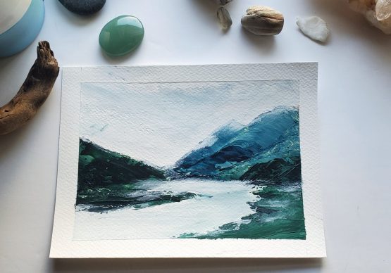 Mist Lake Painting on watercolor paper Blue Mountain range against grey misty skies and grey lake surface below with a dark green forest on the left sitting upon a white table with beautiful green gems and natural stones around the painting