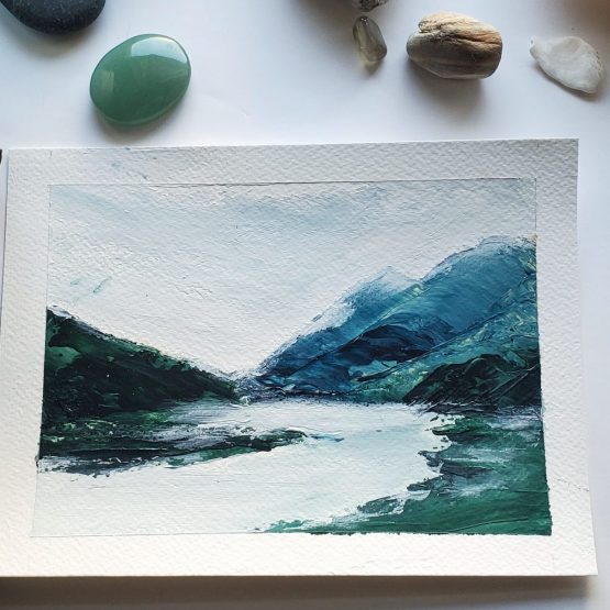 Mist Lake Painting on watercolor paper Blue Mountain range against grey misty skies and grey lake surface below with a dark green forest on the left sitting upon a white table with beautiful green gems and natural stones around the painting