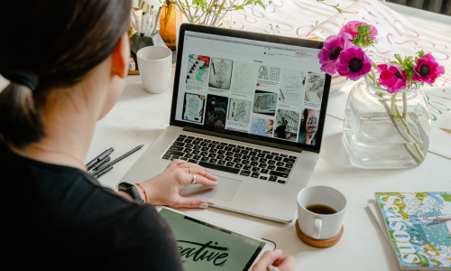 A woman with dark hair pullback in a pony tail sits at a desk in front of her silver laptop writing descriptions for art while her tablet in front of her says Creative. Red flowers sit in a vase next to the silver laptop on the white table with colorful notebooks and pens lay about the table